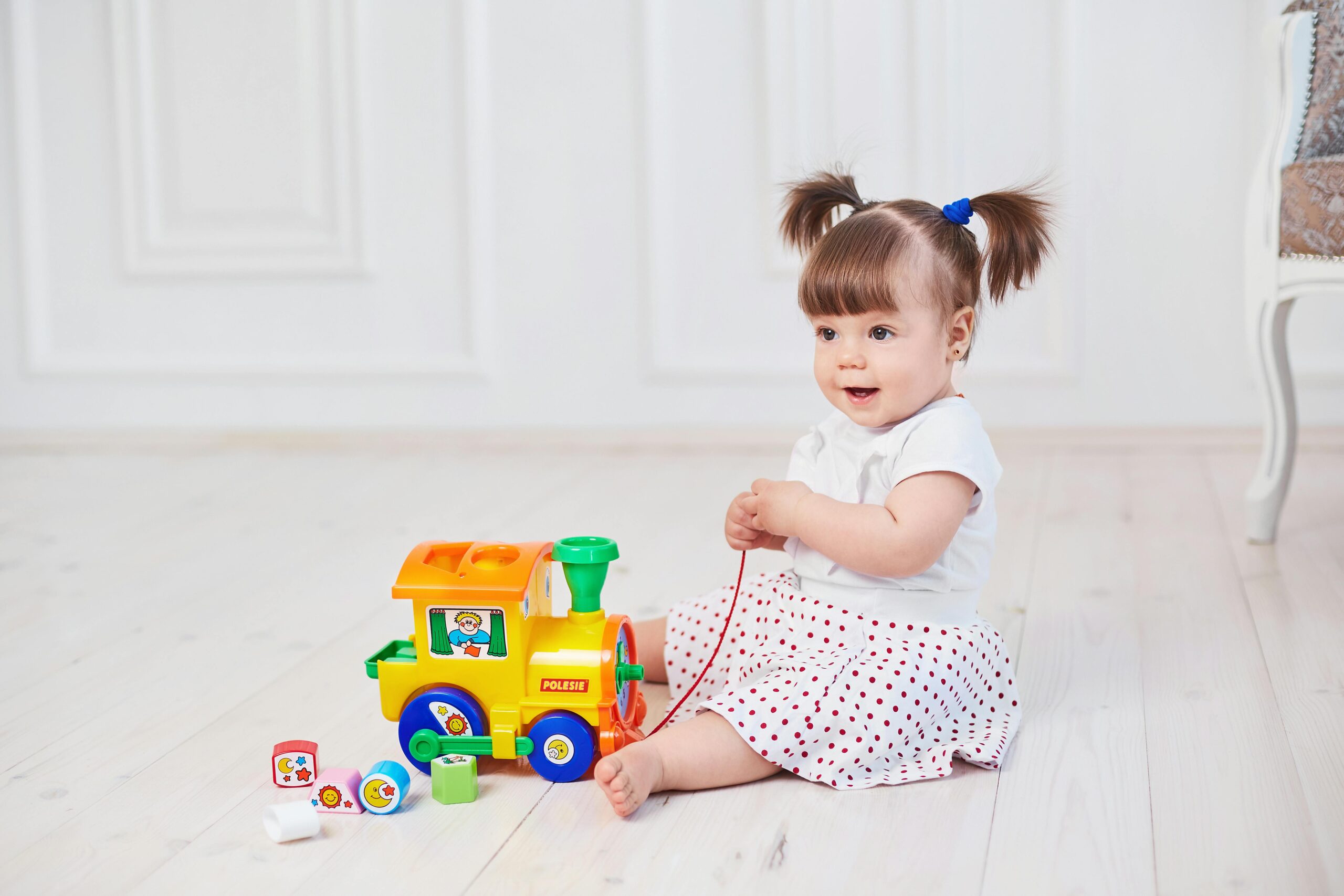 Cute baby girl playing with a bright toy train on a wooden floor indoors.